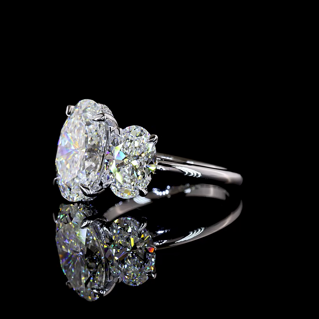 6.15ctw lab grown oval three stone engagement ring in 14k white gold on a black reflective surface.